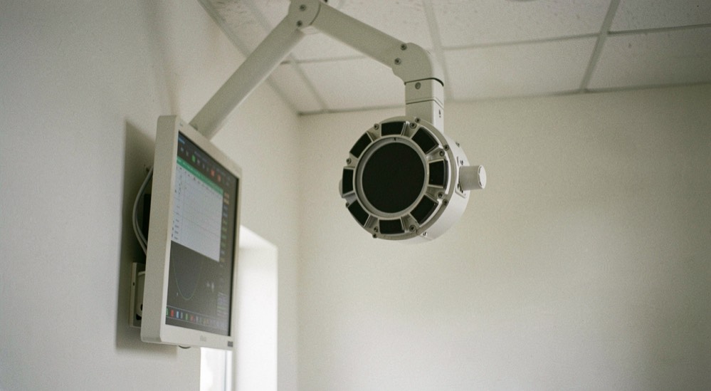 A wall mounted radiography detector panel and a ceiling suspended tube head angled towards it in an otherwise empty room